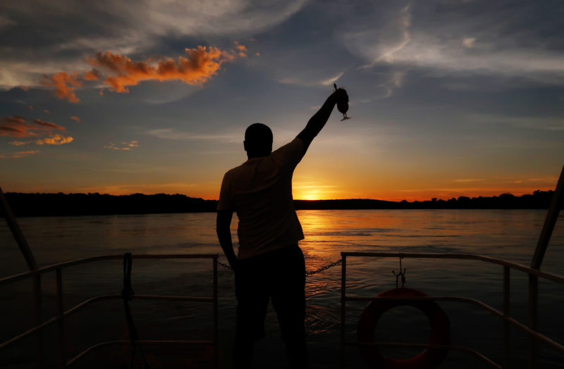 Silhouette of Wildlife photographer Clement Kiragu holding up a cocktail glass on a boat on the Zambezi river in Zambia